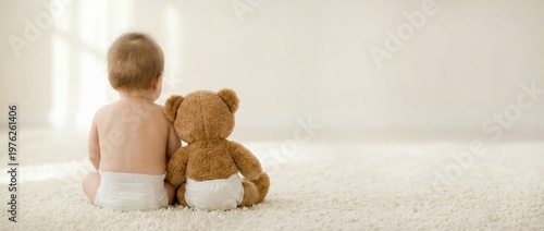 A baby sits on a carpet with a teddy bear both facing away from the camera in a bright room with a window