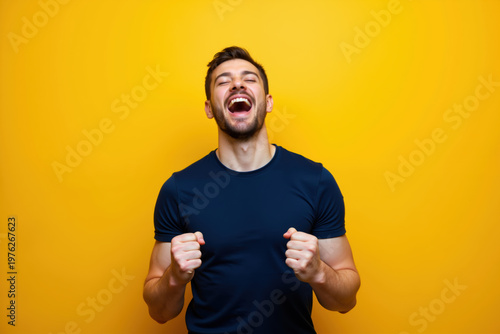 A man wearing a dark blue shirt is laughing heartily with his eyes closed and fists clenched in front of a bright yellow background.