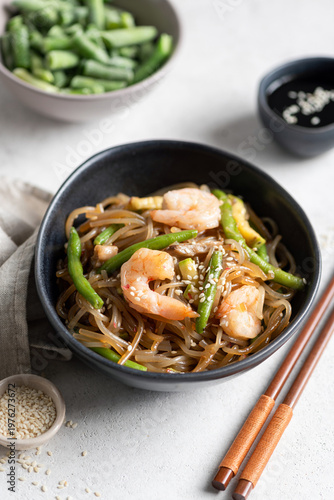 Asian glass noodles with shrimps and green beans served in bowl on grey background, garnished with sesame seeds. Wooden chopsticks on side
