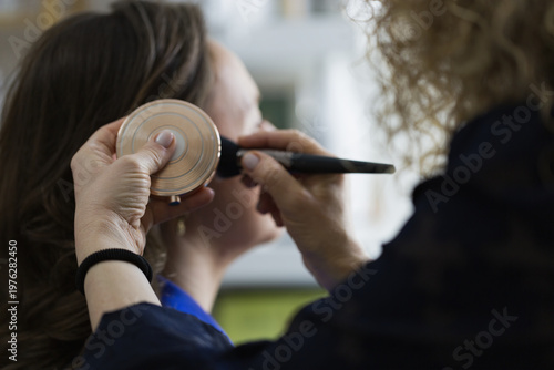 Makeup artist with curly blonde hair applying makeup with brush to young woman who holds golden compact mirror, professional beauty session in studio setting. Make up artist work in beauty salon
