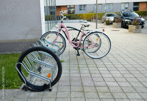 A white bicycle is secured to a metal bike rack on a tiled sidewalk. Colorful artwork adorns the nearby building, creating a vibrant urban atmosphere in the bright daylight. High quality photo