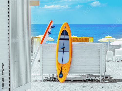 Bright orange SUP board and paddle on a sunny white beach. Summer vacation concept with paddleboard by the turquoise sea. Minimalist aesthetic with copy space.