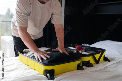 Man closing and packing yellow suitcase on bed at home, preparing for trip or vacation.