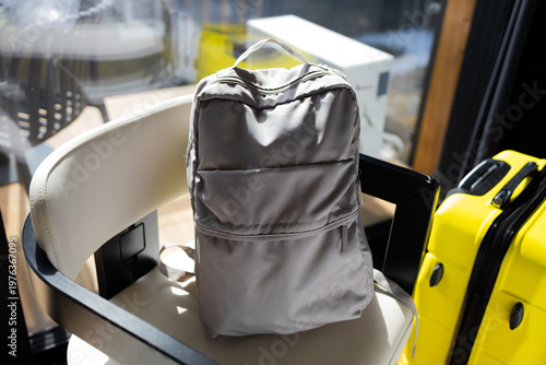 Gray backpack placed on chair next to yellow suitcase near window, travel luggage prepared and ready for trip at home interior.