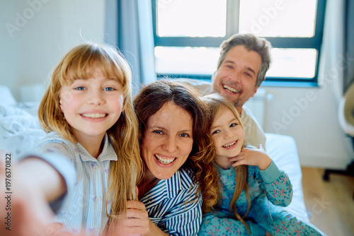 Parents and two daughters enjoy making a selfie in bed, cheerfully smiling, wearing pajamas and bonding in the morning light