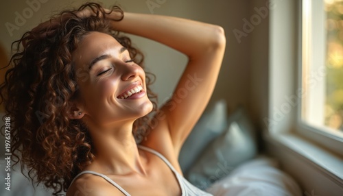 Young woman with curly hair smiles with closed eyes. She stretches arms up indoors near window. Sunlight illuminates her face showing happiness and carefree mood. She feels refreshed and relaxed.