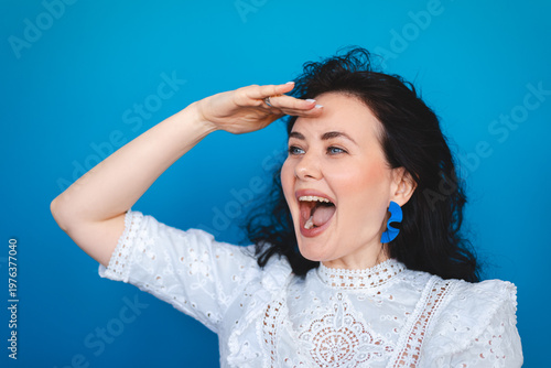 Excited happy woman looking aside discovering amazing opportunities with wide open mouth, smiling girl in white lace dress scouting and searching away for future big sales on blue background