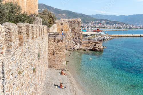 Tersane Beach with clear turquoise water and ancient shipyard walls. A man stands on a wooden viewpoint overlooking the Mediterranean coast. Alanya, Turkey - October 25, 2020