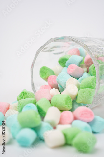 A multitude of soft, pastel-colored mini marshmallows spill from an overturned faceted glass on a clean white surface