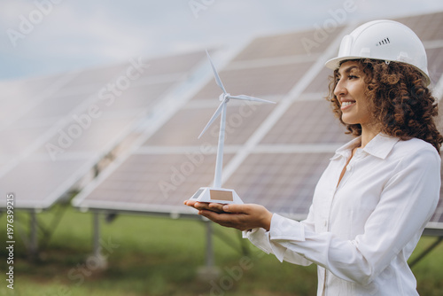 Young woman in hard hat presenting a wind turbine model, standing in front of solar panels, planning green technology