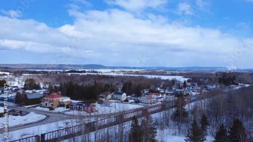 Cinematic drone footage of an empty freight train traveling through the snowy village of Sayabec in the Matapedia Valley, Quebec. A scenic winter landscape with a distant factory in the background.