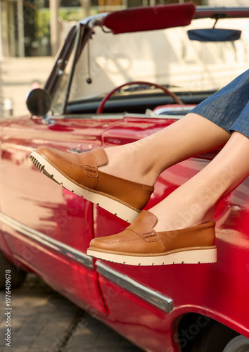 Stylish young woman’s legs wearing tan leather loafers with thick soles, with blue jeans, casually resting on a classic red convertible car, outdoors, showcasing a chic, comfortable, casual style.