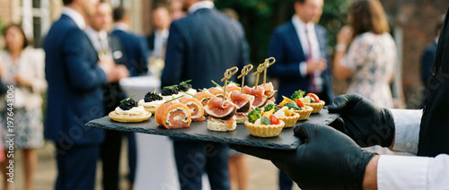 Garçom segurando uma bandeja com canapés e aperitivos durante um evento de catering ao ar livre com convidados ao fundo