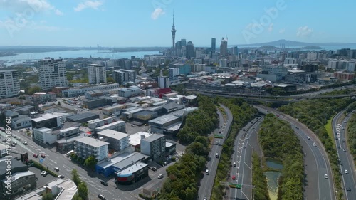 Aerial view of Auckland, New Zealand, showing the city skyline, harbor, and transportation infrastructure. Cars travel on the highway, connecting different parts of the city.