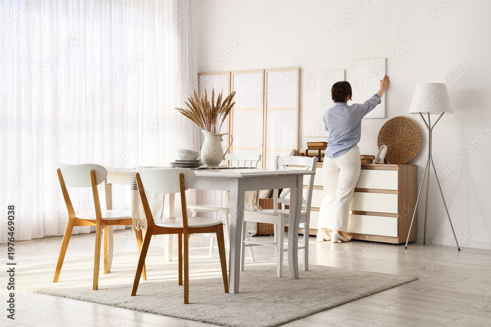 Fototapeta premium Young woman hanging painting on light wall in dining room, back view