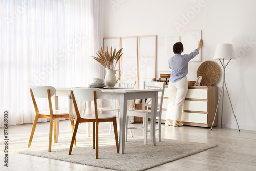 Young woman hanging painting on light wall in dining room, back view
