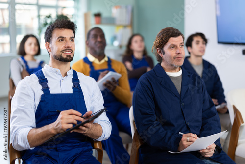 Quadro su tela Young man worker in overalls in multinational group of workers listens to lectur