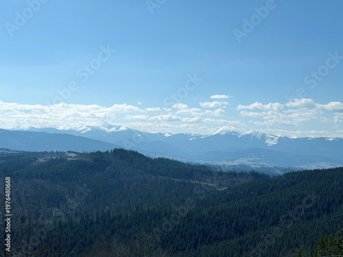 Snow-capped mountain peaks rise above layered blue ridges and dense evergreen forests, creating a serene panoramic landscape ideal for travel, outdoor, nature, background, wallpaper