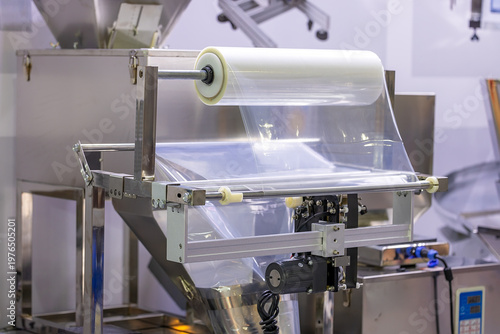 Industrial packaging machine with transparent plastic film roll in a factory setting. Equipment for sealing and wrapping products in manufacturing or food processing industries. Selected focus