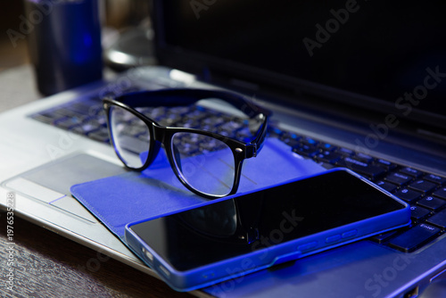 Business closeup portrait in different light with same camera manual and composition. Glasses, mobile phone placed on laptop keyboard. Research, study, work concept in blue lighting