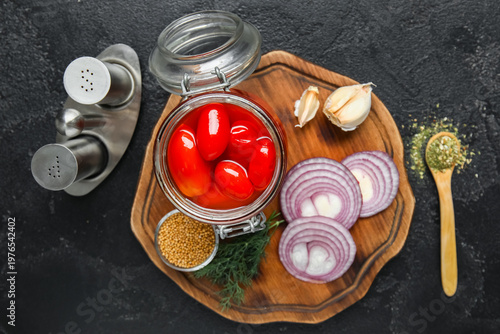 Wooden board with jar of pickled tomatoes and different spices on black background