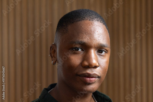 African American man looking at camera before wood-slat wall, dark green shirt, nose-hoop, earrings