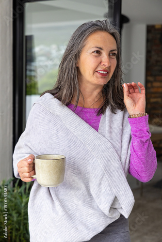Middle-aged woman wearing shawl standing near sliding glass door holding ceramic mug and gesturing