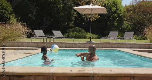 African father and son in swim trunks, youth initiating play, tossing beach ball in backyard pool