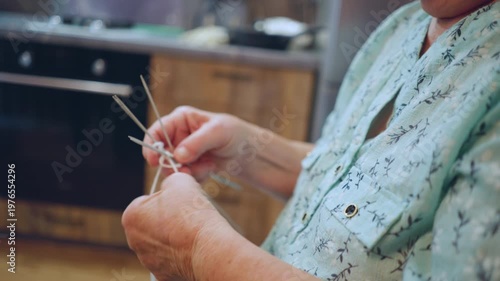 Closeup caucasian hands knitting with needles, kitchen background hints at cozy home routine, knuckles and fingers show practiced dexterity while wool loops form intricate stitch work with gentle