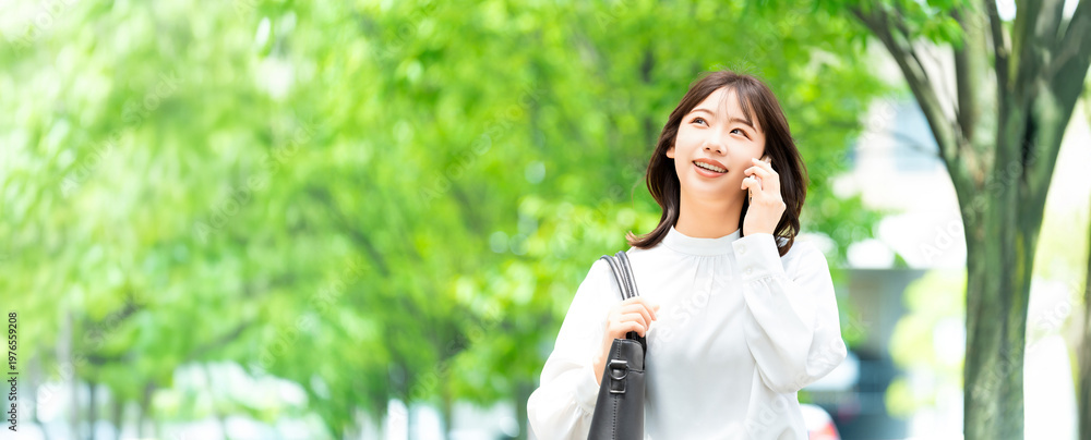 Fototapeta premium 遊歩道でスマートフォンで話す日本人女性・ビジネスウーマンのビジネスイメージ(LP・バナー向け), Japanese Businesswoman Talking on Smartphone on Promenade for Landing Page Banner