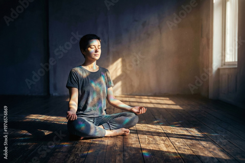 A young woman meditating on a wooden floor in a dark empty room illuminated by window sunlight and rainbow prism reflections