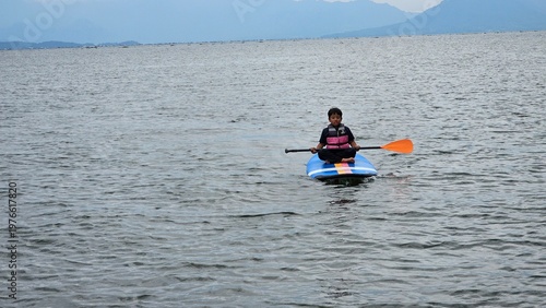 An East Asian boy is sitting on stand up paddle board at a beautiful lake under blue sky on with amazing hills and mountain in background.