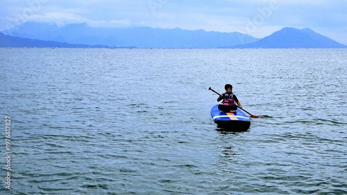 An East Asian boy is sitting on stand up paddle board at a beautiful lake under blue sky on with amazing hills and mountain in background.