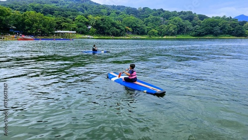 Exited East Asian kids are sitting on stand up paddle board at a beautiful lake under blue sky on with amazing hills and mountain in background. 