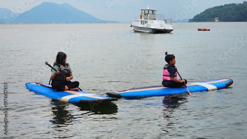 Exited East Asian kids are sitting on stand up paddle board at a beautiful lake under blue sky on with amazing hills and mountain in background. 