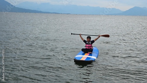 An East Asian boy is sitting on stand up paddle board at a beautiful lake under blue sky on with amazing hills and mountain in background.