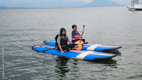 Exited East Asian kids are sitting on stand up paddle board at a beautiful lake under blue sky on with amazing hills and mountain in background. 