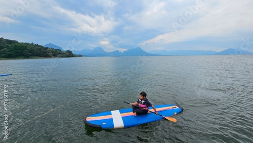 An East Asian boy is sitting on stand up paddle board at a beautiful lake under blue sky on with amazing hills and mountain in background.