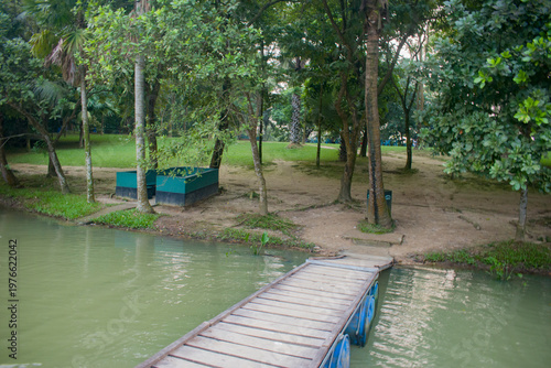 A wooden dock leading to a calm park pond surrounded by trees and lush greenery.