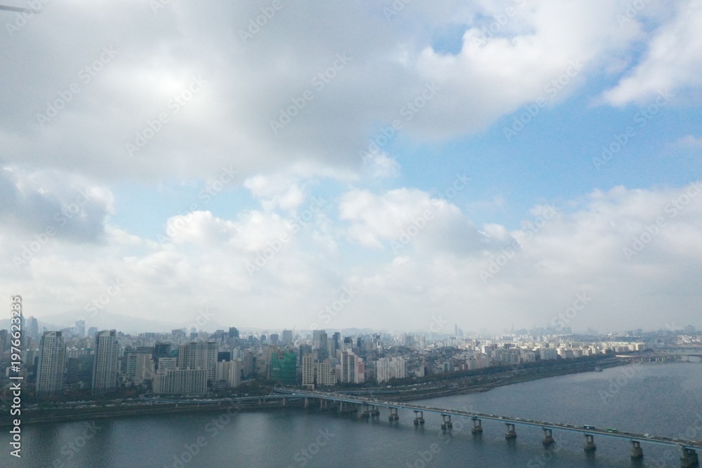 Obraz premium Wide aerial shot of a city skyline beside a river under a soft cloudy sky.