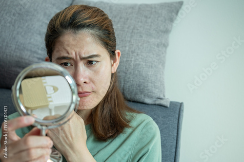 Asian woman worried about her face when she saw acne and freckles occur on her face by mini mirror.