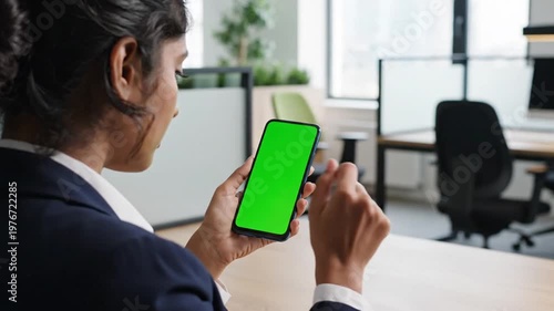 A woman in a business suit holds a smartphone with a green screen in an office setting, interacting with the device.