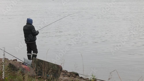A man standing on the river bank. Fisher in a jacket with a fishing rod stands on the river bank. An angler with a spinning rod on the river bank catches fish. Fisherman in nature.