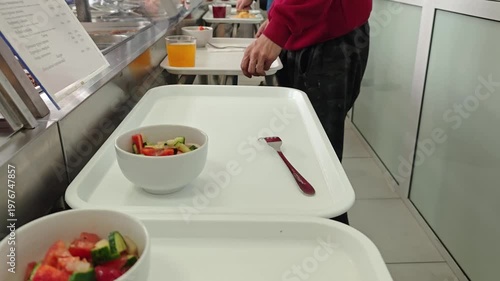 A person carrying a white tray with a fresh vegetable salad and a fork in a cafeteria or dining hall setting.