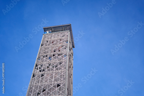A modern observation tower with a distinctive geometric lattice facade made of crisscrossing wooden or metal slats forming triangular patterns, photographed from a low angle against a clear blue