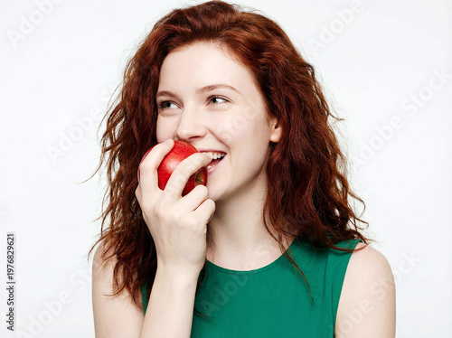 Redhead woman biting a fresh red apple and smiling against a white background.