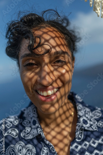Cheerful Indian woman sporting beautiful, curly hair beams with a bright smile, her face beautifully adorned with intricate shadow patterns, all set against the backdrop of a serene, clear blue sky