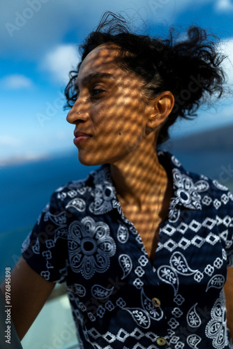 Profile of a beautiful indian woman with curly hair, wearing a vibrant patterned shirt. Intriguing shadows from a nearby structure enhance the depth and artistic flair of the scene
