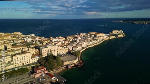Aerial view of Ortigia Island showcasing historic architecture, coastal waters, and vibrant city life, with boats docked along the shoreline and lush greenery in the foreground