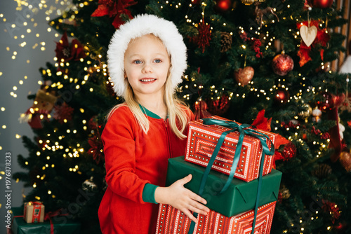 Merry Christmas and Happy Holidays. Little child under Christmas tree. baby girl in Santa Claus hat with gifts under Christmas tree with many gift boxes presents. Happy Holidays, New year.